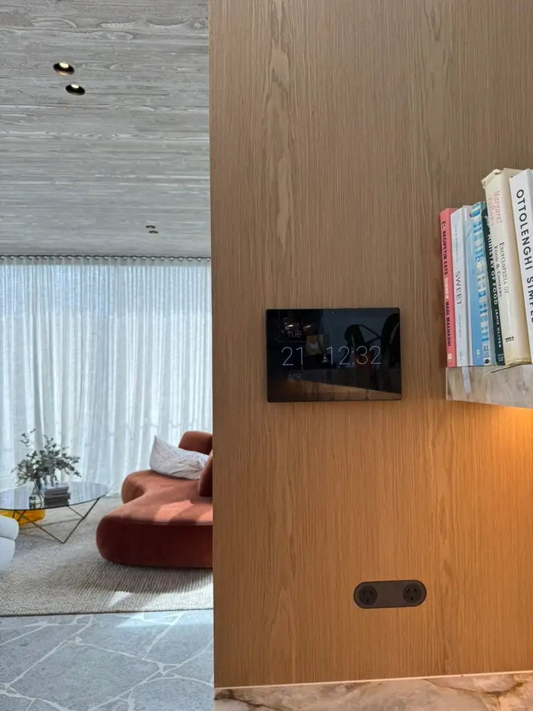 Wall-mounted screen set into timber joinery beside books and a living room with exposed concrete ceiling